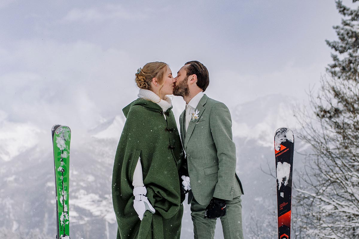 A couple dressed in formal attire kisses outdoors in a snowy Austrian mountain setting, standing between two pairs of skis—a magical scene perfect for wedding videography.