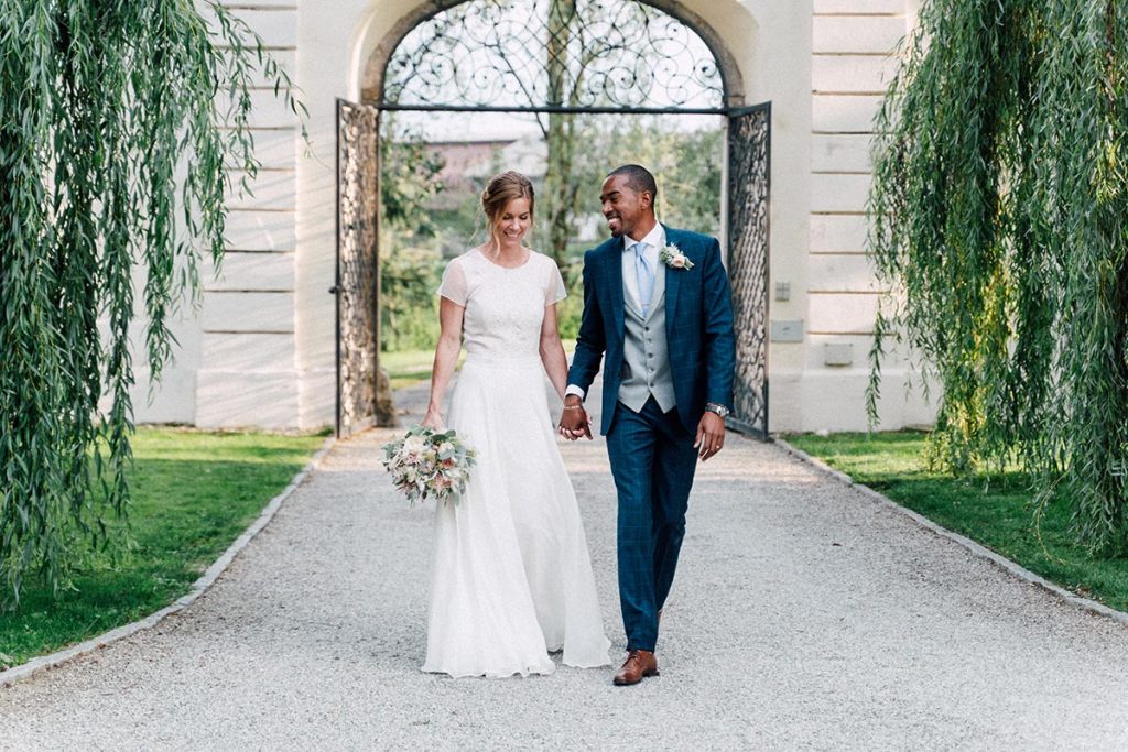 A bride and groom walk hand in hand on a gravel path outside, surrounded by greenery and an arched gate in the background—an enchanting scene perfect for timeless wedding videography in Austria.