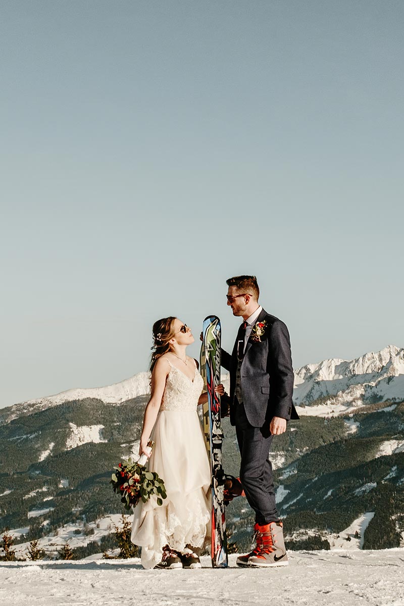 A bride and groom stand on snow with ski gear, dressed in wedding attire and holding bouquets, as majestic Austrian mountains rise in the background under a clear sky