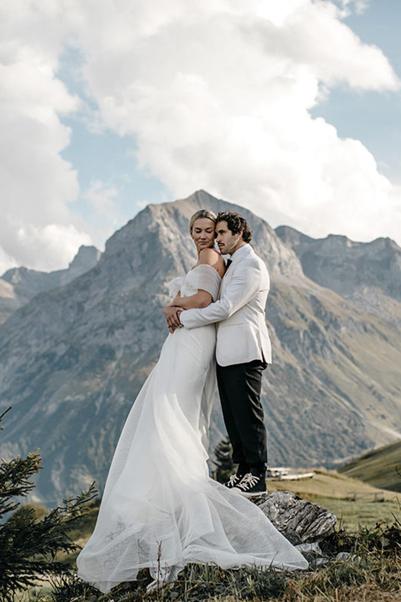 A bride and groom in formal attire stand embracing on a grassy hilltop with mountains and clouds in the background, captured beautifully by an expert Alps wedding videographer Story Of Your Day