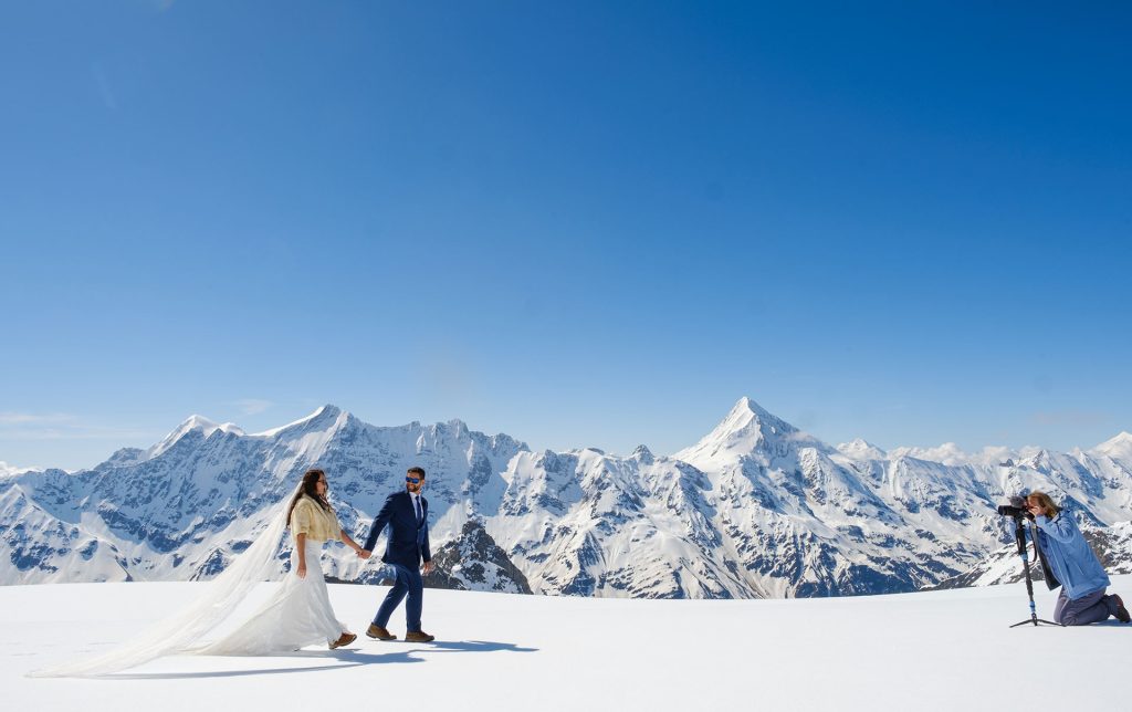 A bride and groom walk hand in hand on snow with mountains in the background while a Emma Wilson from Story Of Your Day films them.
