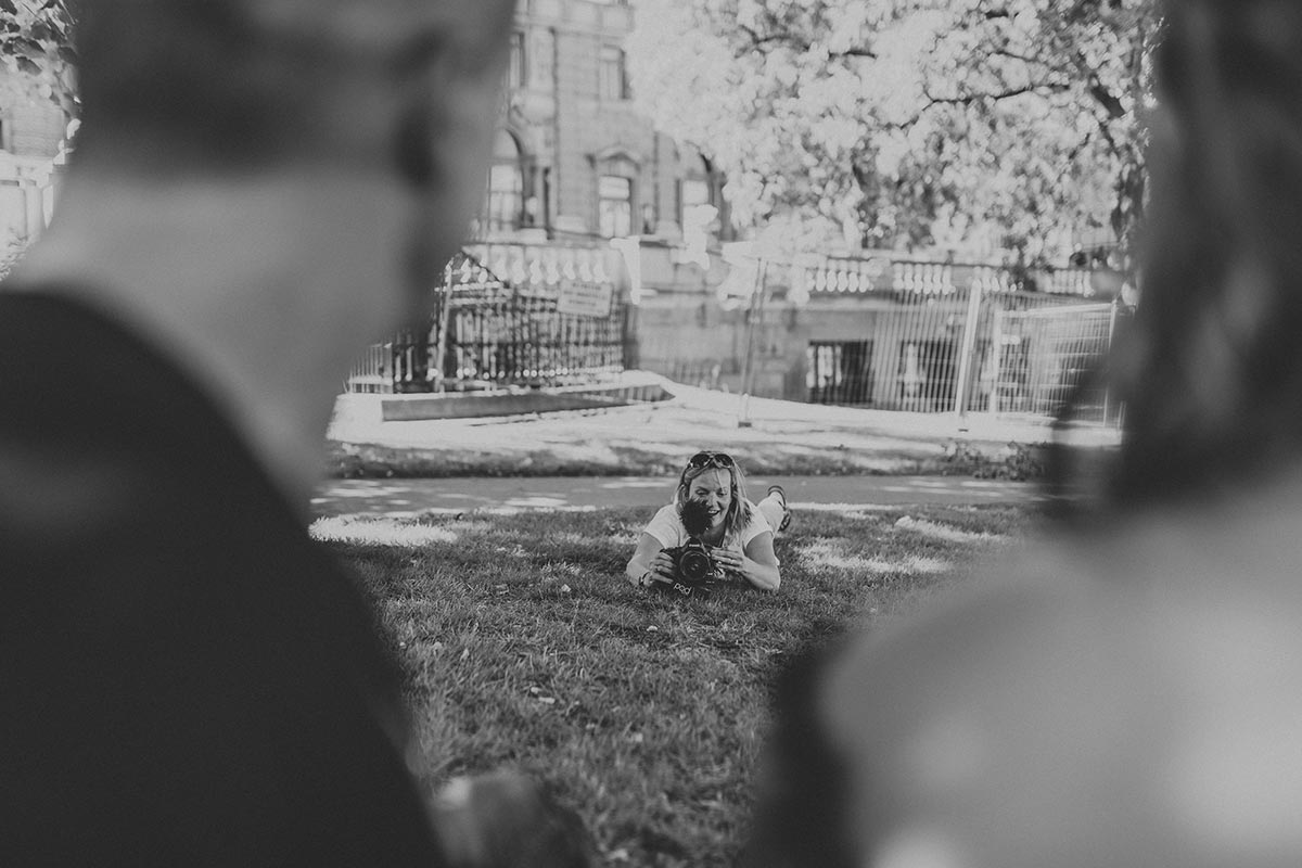 Emma Wilson lies on grass aiming a camera at two blurred people in the foreground, with trees and a building visible in the background.