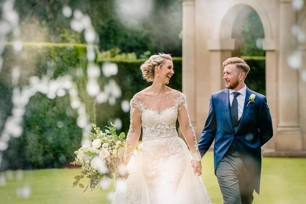 A bride and groom walk hand in hand outdoors, smiling at each other, with greenery and an arched stone structure in the background—perfectly captured by a UK Wedding Videographer.
