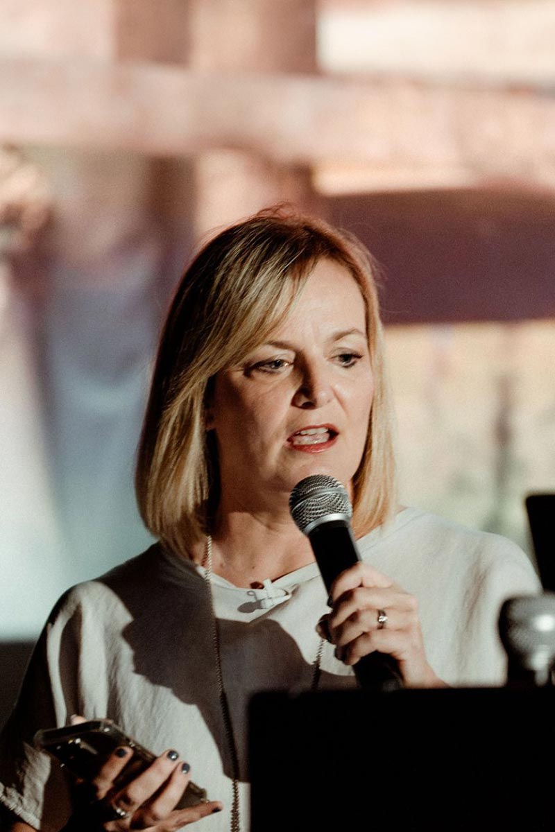 Emma Wilson is speaking into a microphone while holding a phone, standing indoors with a blurred media background.