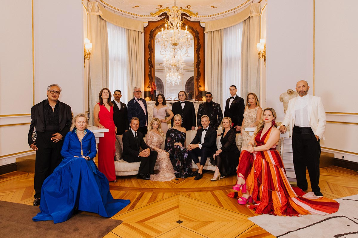 A group of people dressed in formal evening wear pose together in an elegant, chandelier-lit room with ornate decor and wooden floors, perhaps gathered for a media event or speaking engagement.