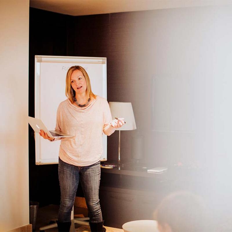 A woman stands in front of a flip chart, holding a laptop and a marker, speaking confidently as she gives a media presentation in an office or meeting room.