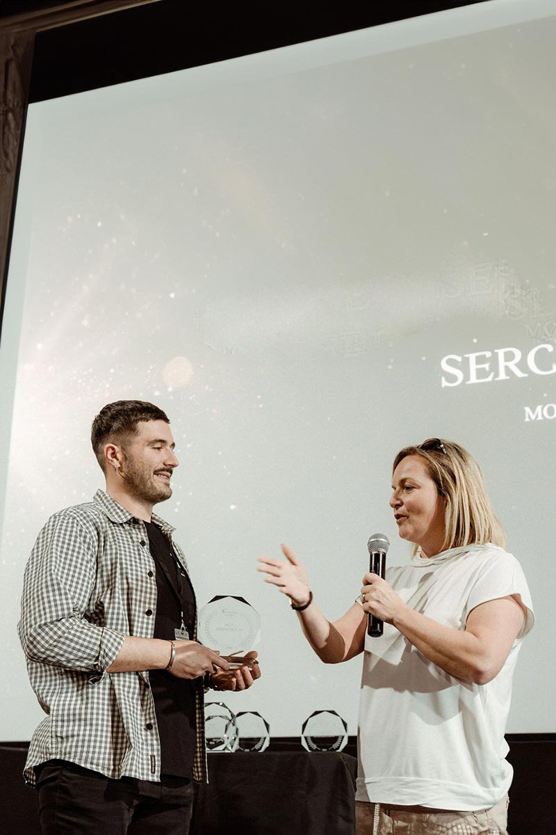 A man receives an award from Emma Wilson holding a microphone on stage, engaging in public speaking, with a projected media screen in the background.