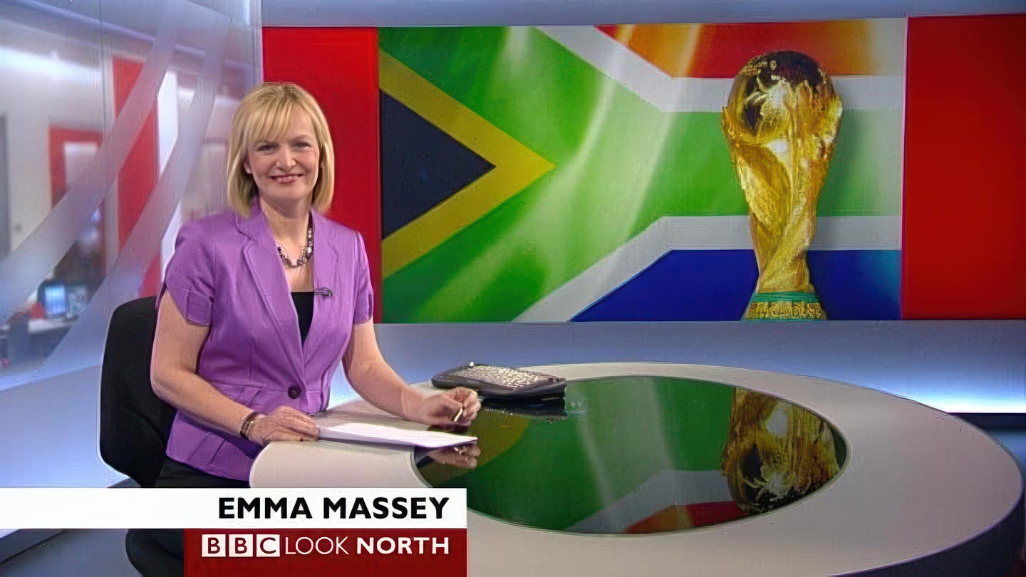 A news presenter is speaking at a desk with a graphic of the South African flag and the FIFA World Cup trophy on the screen behind her. On-screen text reads "Emma Massey, BBC Look North," highlighting her role in media coverage.