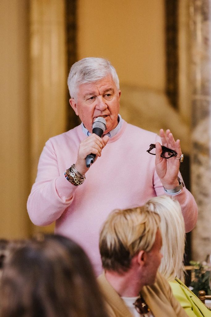 Johnny Roxburgh in a pink sweater speaks into a microphone while holding eyeglasses, talks with a seated group of people indoors.