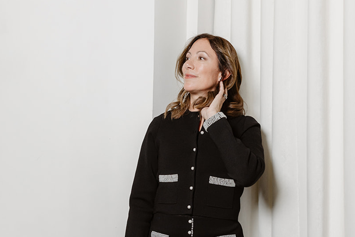 Savina Uldry stands indoors, wearing a black jacket with silver trim, looking to the side with one hand near her ear. White curtains hang in the background.