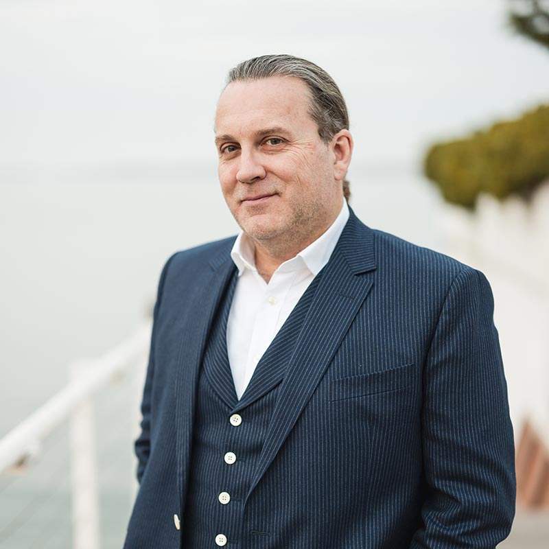 James Lord, dressed in a blue pinstripe suit and white shirt, stands outdoors near a railing, with water and greenery in the background.