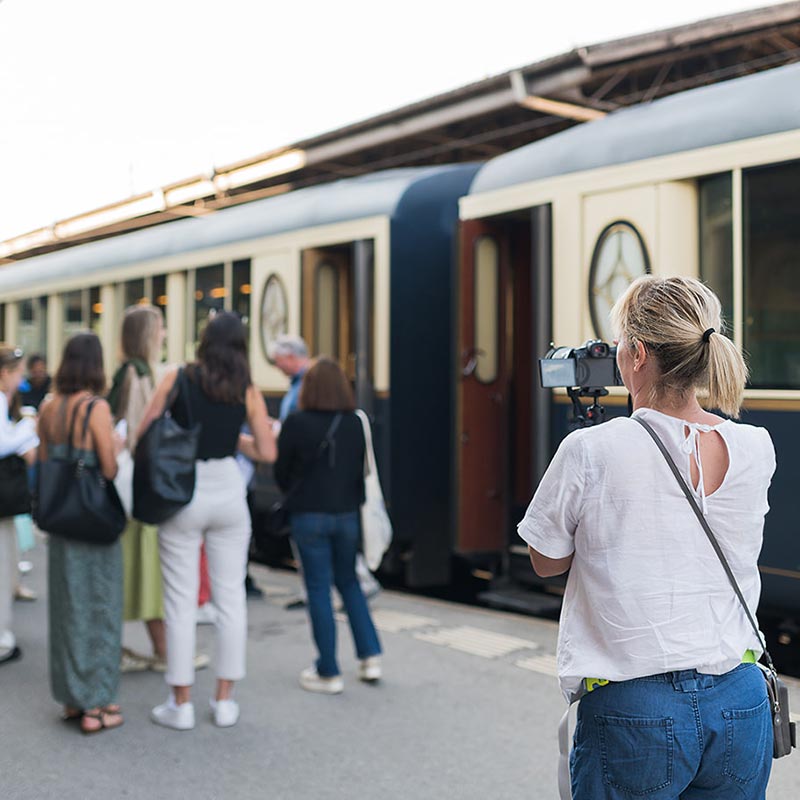 A group of people waits outside a vintage-style train, while a woman in the foreground captures the story of your day with her camera.