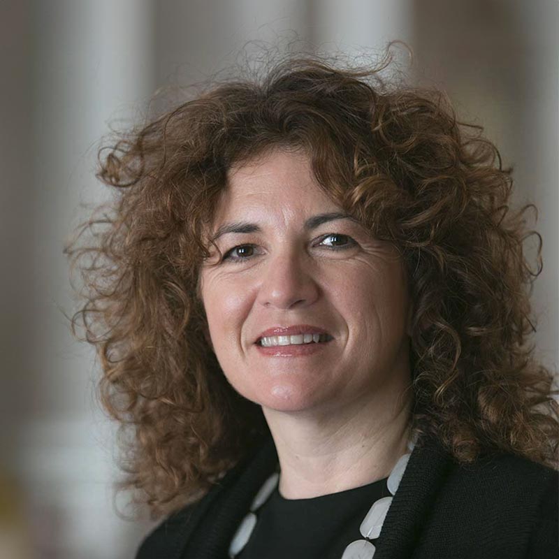 Sonia Petrazzi, a woman with curly brown hair, smiles at the camera, wearing a black top and a chunky necklace, with a blurred neutral background.