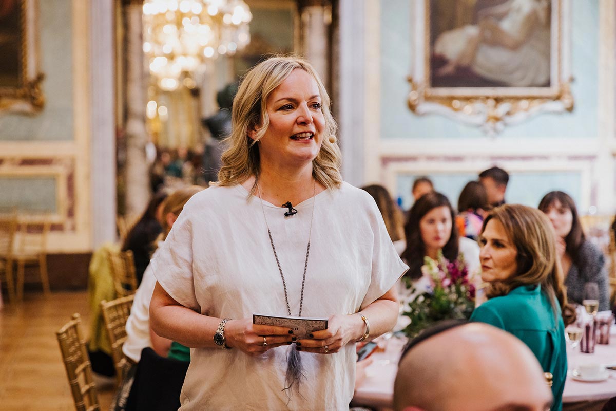Emma Wilson stands and speaks to a seated group in an ornate room with chandeliers and framed paintings, holding a card and wearing a microphone, as she shares the story of your day.