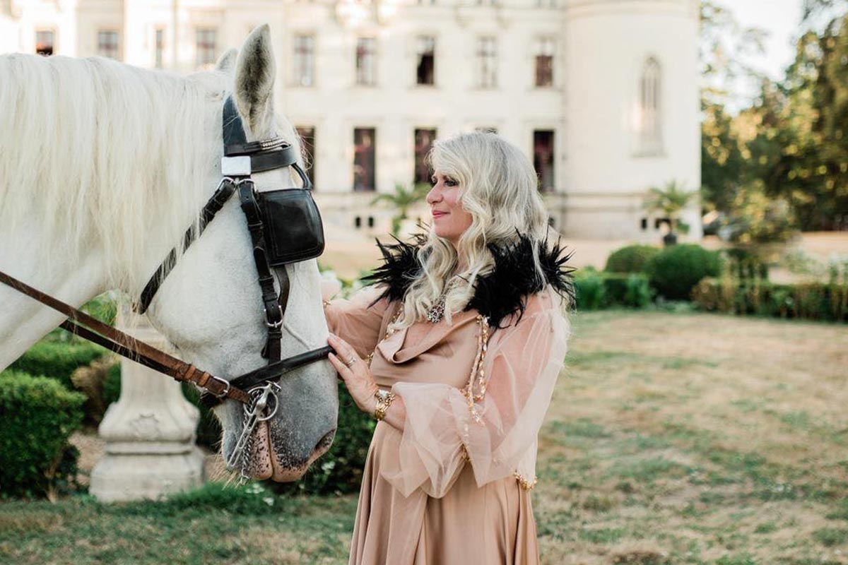 Cynthia Nicholson, in a peach dress with black feather accents, stands outdoors, gently touching a white horse's face near a large white building.