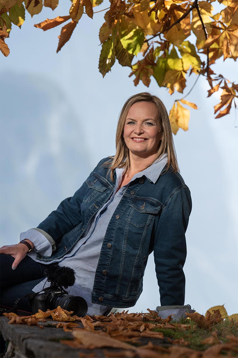 Emma Wilson in a denim jacket sits outdoors on a stone ledge covered with autumn leaves, holding a camera with a microphone beneath tree branches.