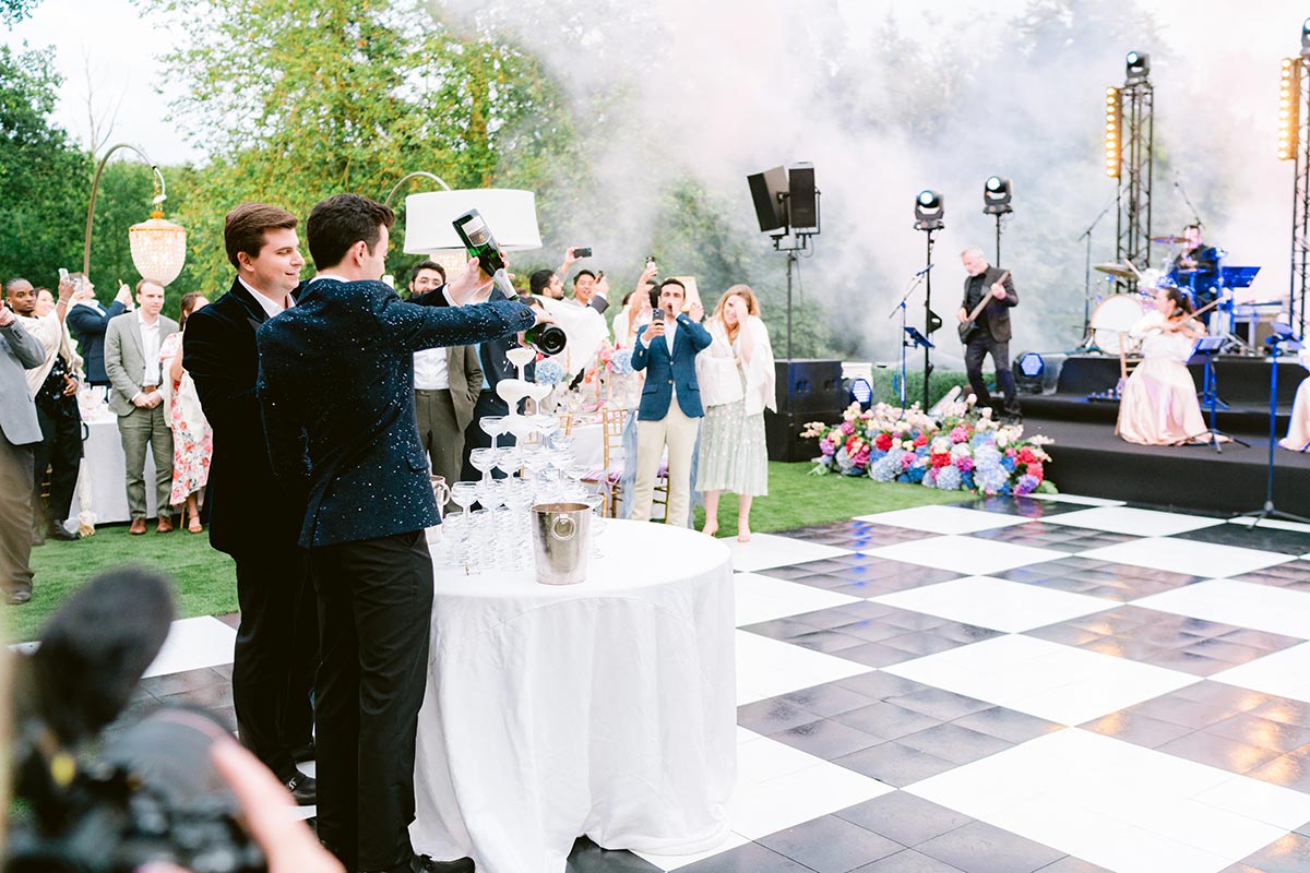 Two grooms pour champagne into a tower of glasses at their wedding with guests watching, a black-and-white dance floor, and a live band—capturing the joy and elegance of inclusive weddings.
