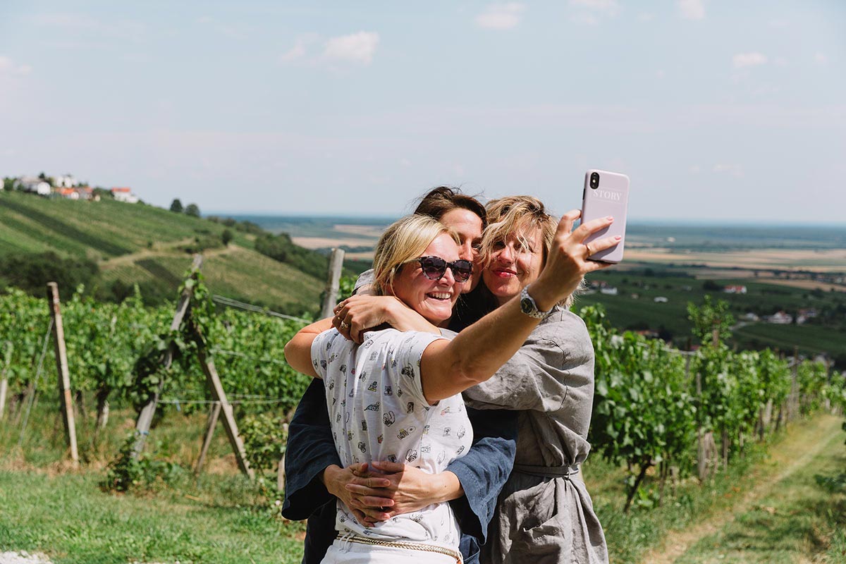 Two brides and their videographer Emma Wilson stand close together, smiling and taking a selfie in a vineyard with hills and a distant view, celebrating love at an LGBTQ+ inclusive wedding.