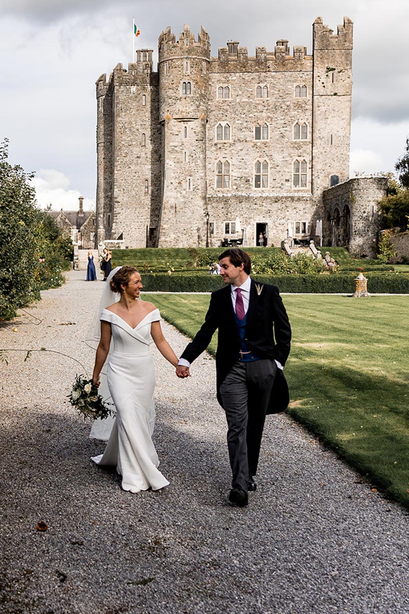 A bride and groom walk hand in hand outside a large stone castle, with manicured lawns and a few people in the background—an idyllic moment captured by a leading UK Wedding Videographer.