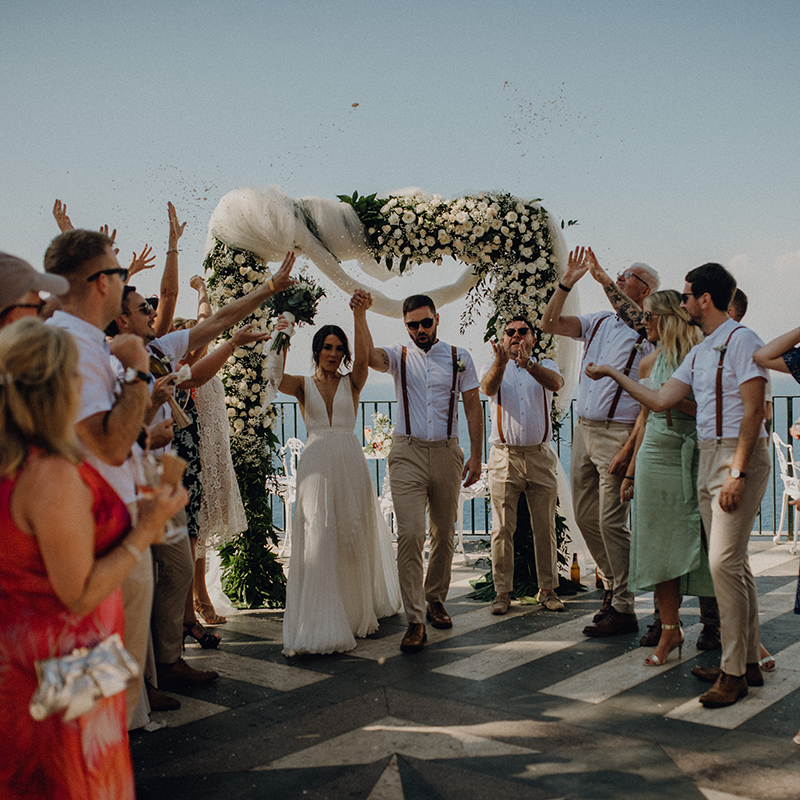 A bride and groom walk under a floral arch as guests celebrate around them outdoors, with confetti thrown in the air—captured perfectly for their Italy wedding video.