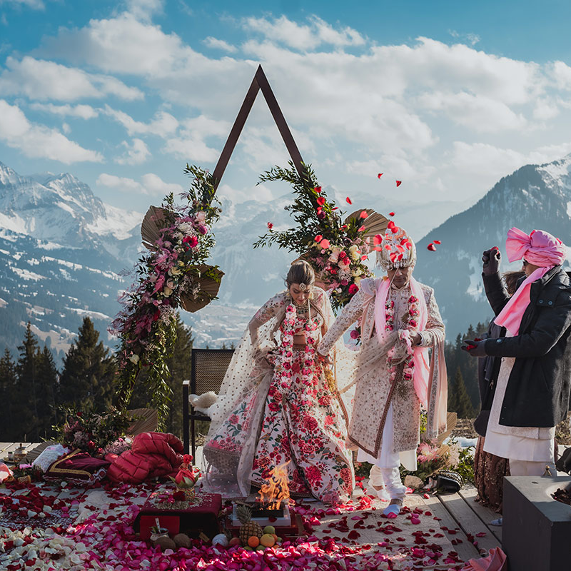 A couple in traditional Indian wedding attire stands under a decorated arch outdoors, with mountains in the background and flower petals scattered on the ground—a magical Switzerland wedding perfect for stunning wedding videography.