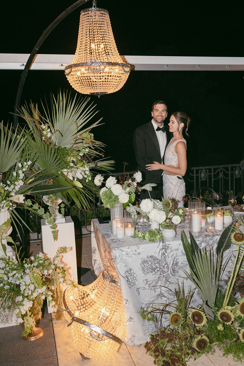 A couple dressed in formal attire stands behind a decorated outdoor table with floral arrangements and two chandeliers, creating a picturesque scene perfect for Switzerland wedding videography.