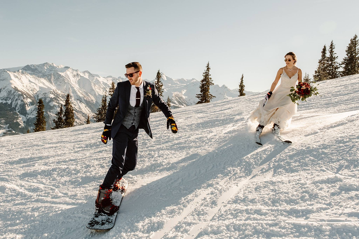 A bride and groom in wedding attire snowboard down a snowy mountain slope in Switzerland, surrounded by evergreen trees and distant peaks—perfect memories for wedding videography.