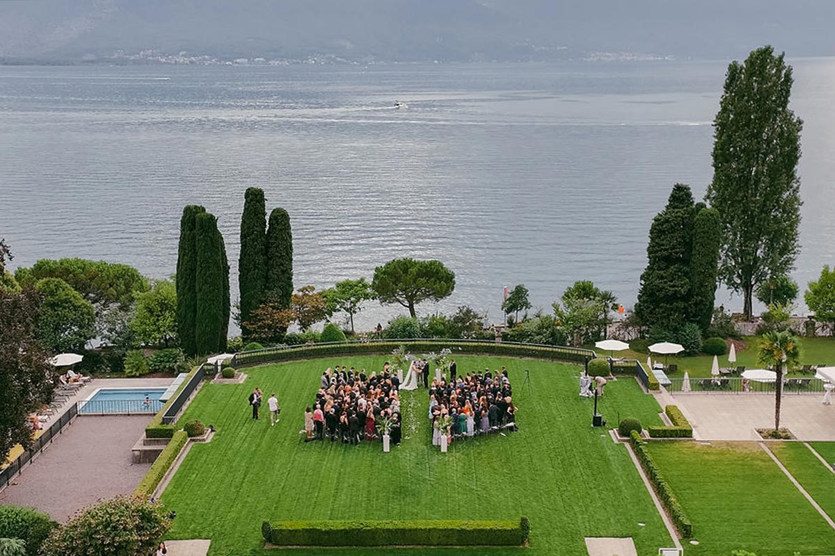 A group of people are gathered on a large, manicured lawn by a lake in Switzerland, with trees and landscaped gardens surrounding the area—an ideal setting for stunning wedding videography.