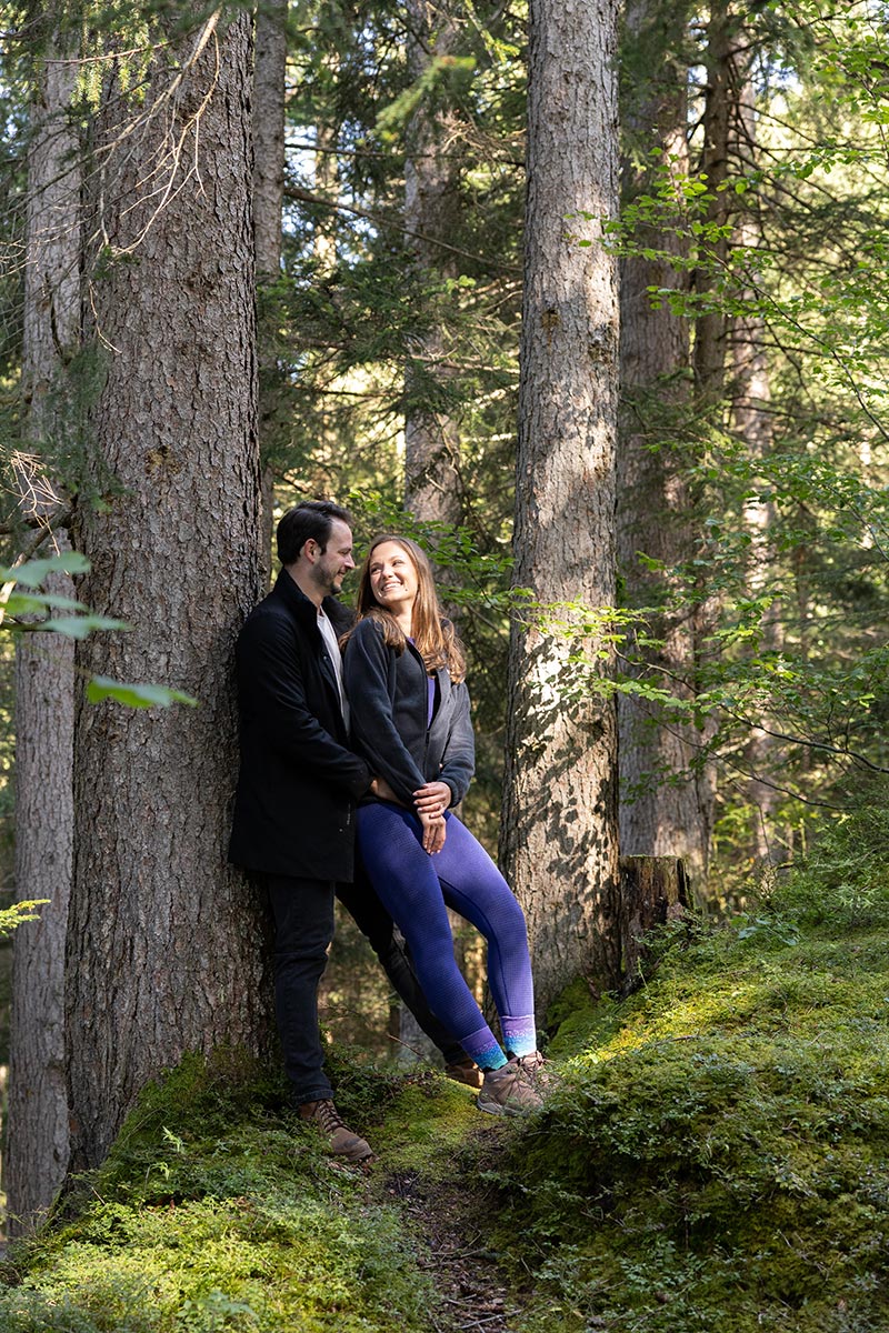 Deux personnes se tiennent près l'une de l'autre dans une forêt ensoleillée, appuyées contre un arbre et se souriant, entourées de verdure et de grands arbres. Elles profitent de la beauté de la nature avec la tranquillité d'esprit que procure l'assistance de luxe offerte par Swiss Concierge.