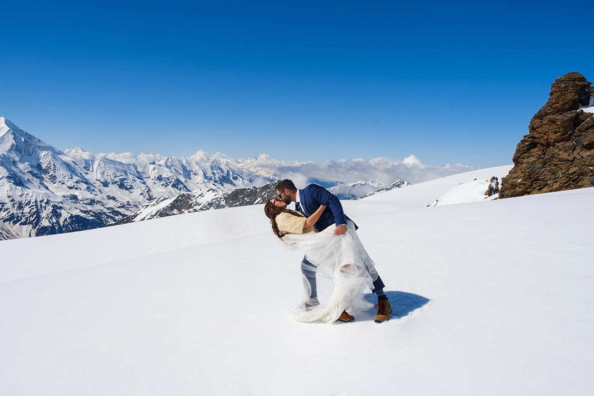 Un couple en tenue de mariage s'embrasse dans un paysage montagneux enneigé, sous un ciel bleu clair et avec des sommets enneigés au loin, profitant de la magie organisée par un concierge de luxe en Suisse.