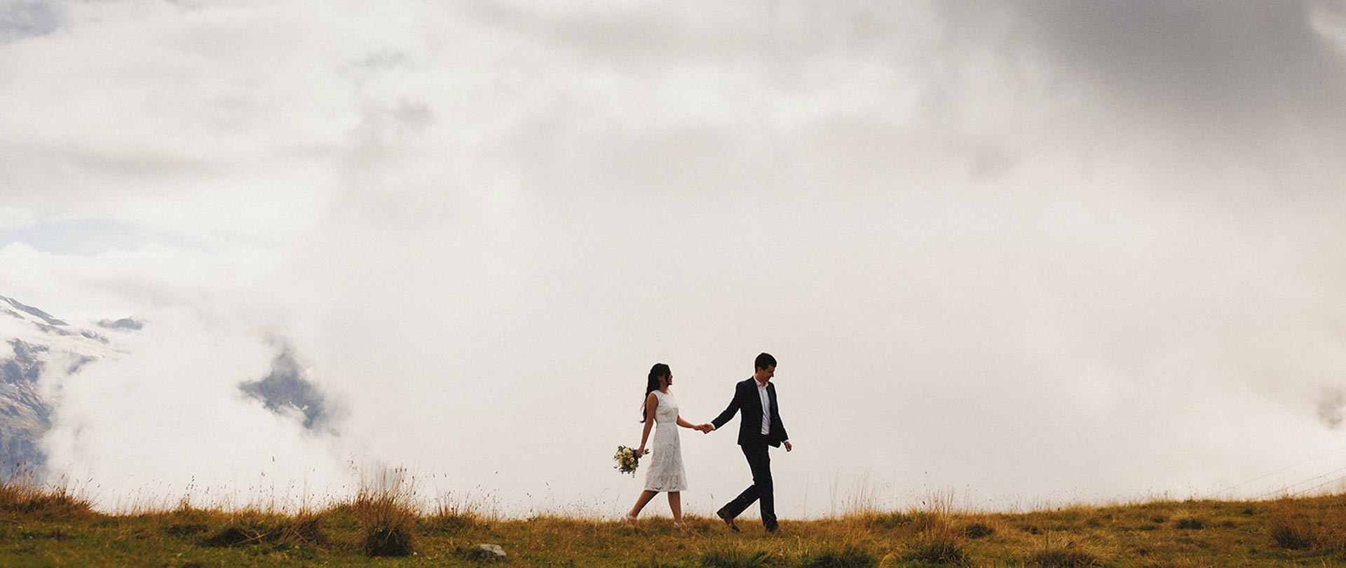 A couple dressed in formal attire walks hand in hand on a grassy hill under a cloudy sky, enjoying the bespoke service of a Swiss Concierge.