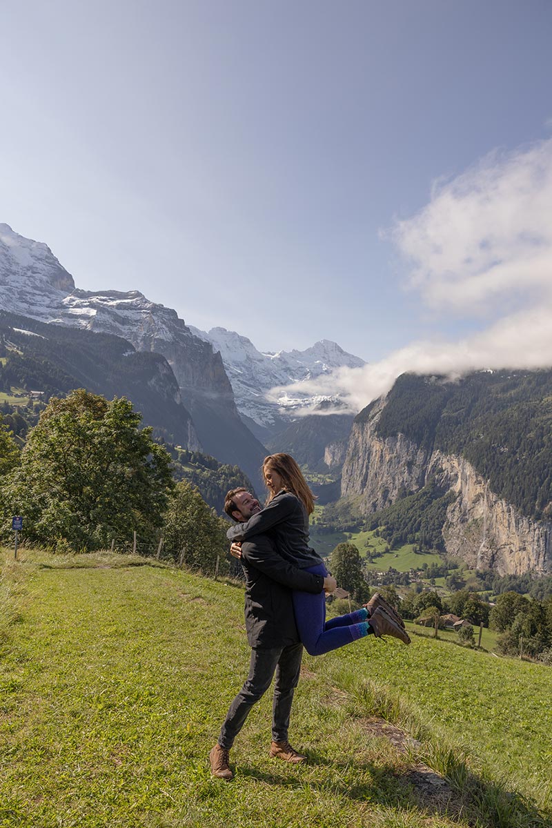 Un homme soulève une femme dans ses bras alors qu'ils sourient, debout sur l'herbe avec des montagnes et une vallée en arrière-plan, sous un ciel partiellement nuageux en Suisse, lors de leur demande en mariage à Wengen.