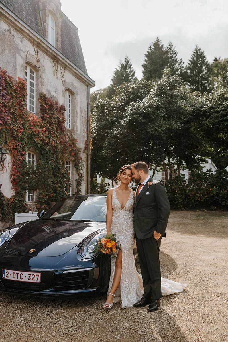 A bride and groom stand together beside a black convertible sports car in front of a large ivy-covered building in France, holding a bouquet