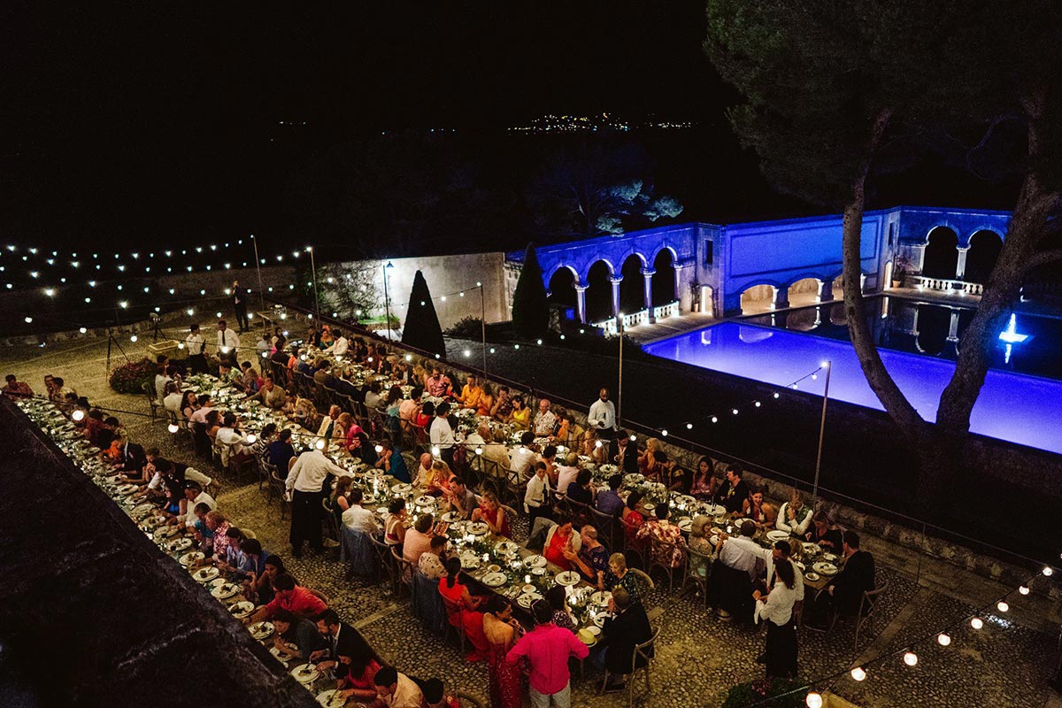 An outdoor evening banquet with long tables of guests dining beside a pool, string lights overhead, and waiters serving food—perfect for a destination wedding and capturing unforgettable moments on your wedding video.