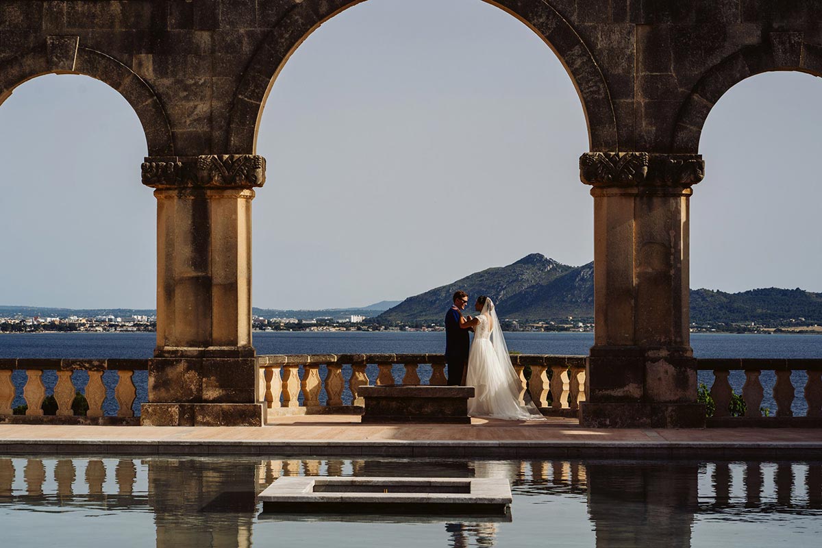 A bride and groom stand together under a stone archway by a pool, with a lake and mountains in the background