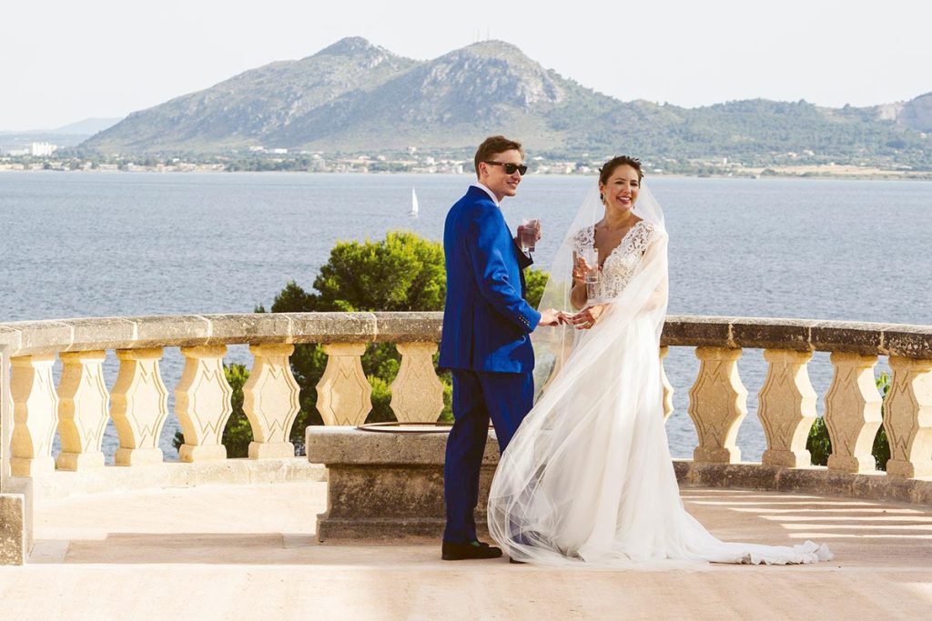 A bride and groom stand together on a terrace overlooking water and mountains, with the groom in a blue suit and the bride in white