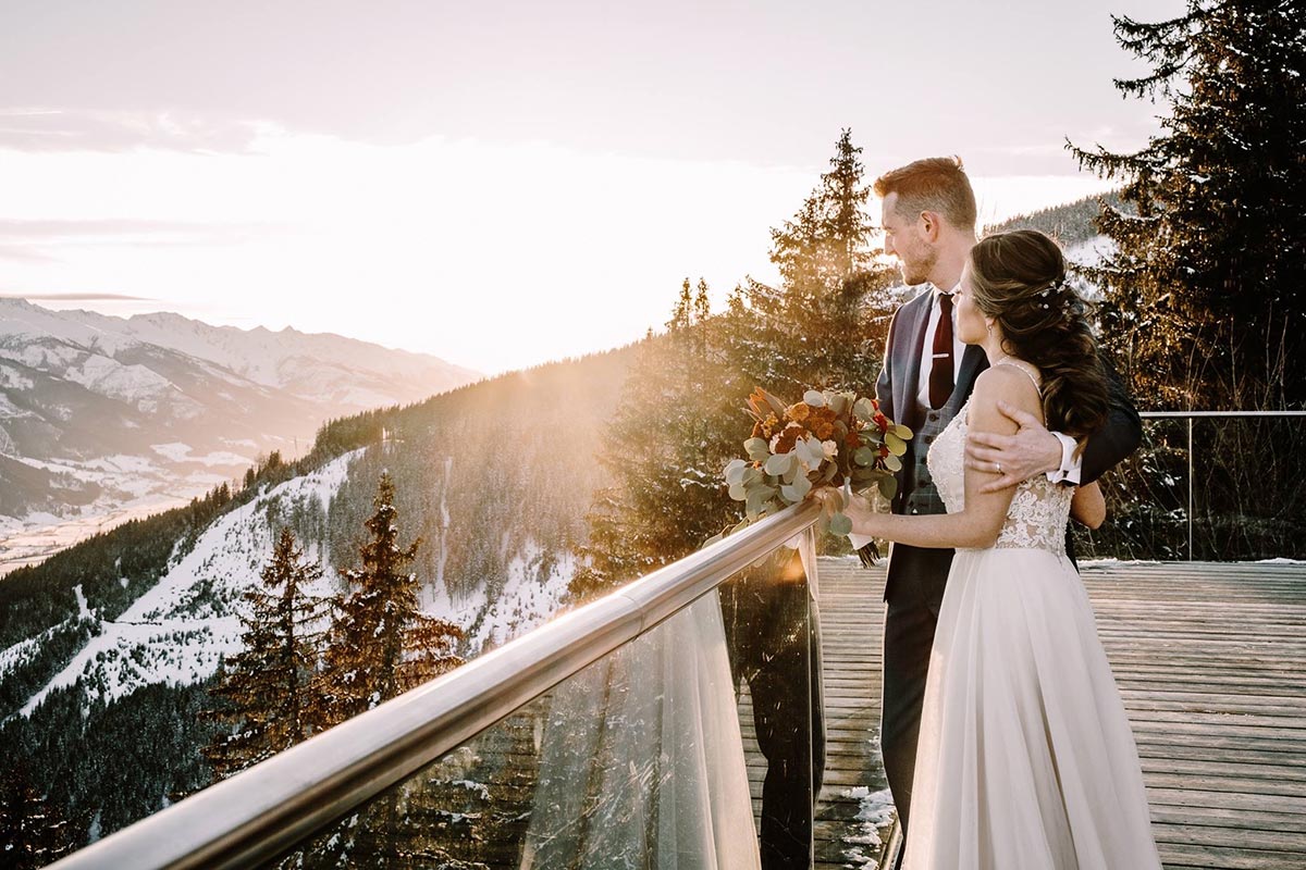 A bride and groom stand together on a wooden deck overlooking a snowy mountain landscape at sunset in Austria, with the bride holding a bouquet