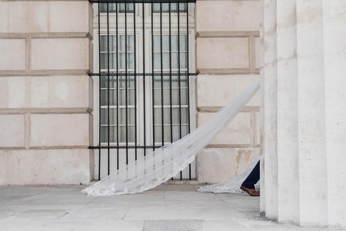 A long white wedding veil trails behind a person partially hidden behind a large stone column in front of a barred window, evoking the elegance captured by top Wedding Videography in Austria.