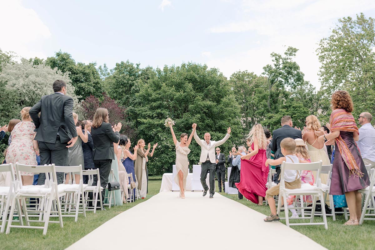 A bride and groom walk down an outdoor aisle with arms raised as guests on both sides stand and cheer, beautifully captured in this Austria wedding video.