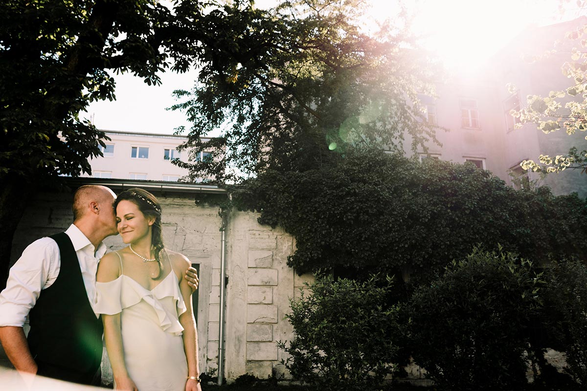 A couple stands close together outdoors near a building covered in greenery in Austria, with sunlight streaming through the trees—a beautiful setting perfect for wedding videography.