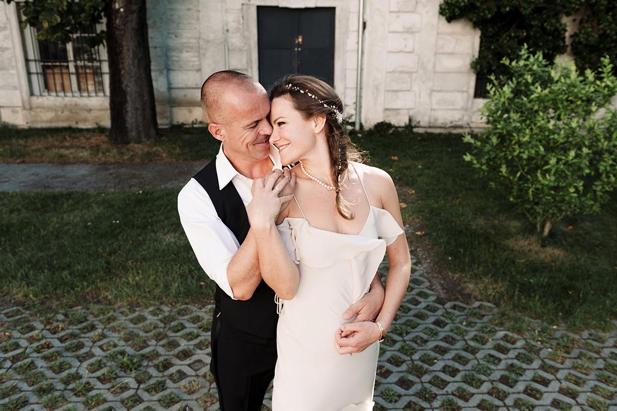 A couple in wedding attire stands close together outside, smiling and embracing in front of a stone building with greenery— a perfect moment captured for a timeless wedding video in beautiful Austria.