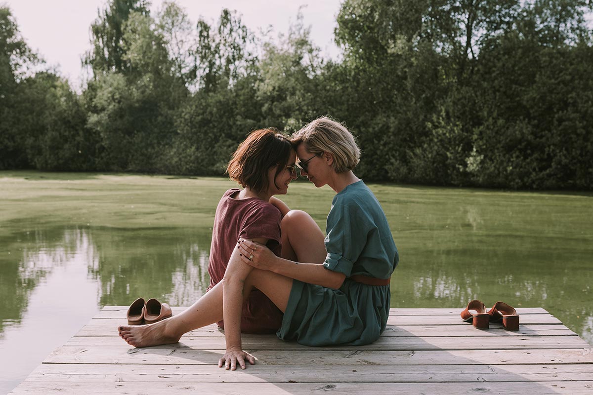 Two brides sit closely together on a wooden dock by a calm, greenish lake in Austria, facing each other with foreheads touching. Shoes are placed beside them. Trees are visible in the background