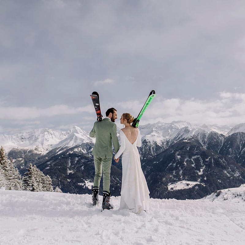 A bride and groom stand looking over the mountains holding their skis during their Swiss Alps adventure elopement