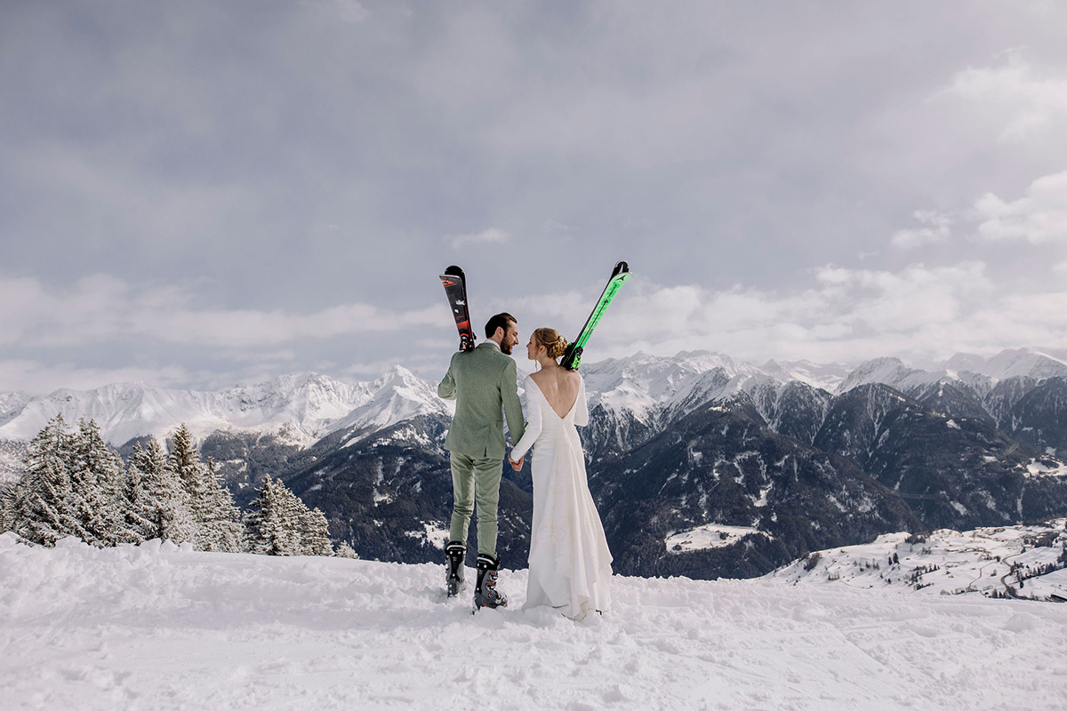 A bride and groom in their wedding outfits look over a snowy valley while holding their skis on their adventure elopement