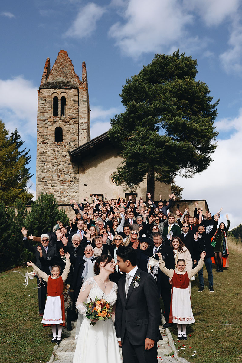 A bride and groom kiss in front of the church with all their guests celebrating behind them