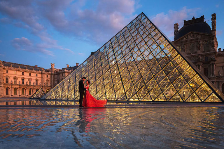 A bride in a red dress embraces her groom in from of the glass pyramid of the Louvre on their wedding in Paris