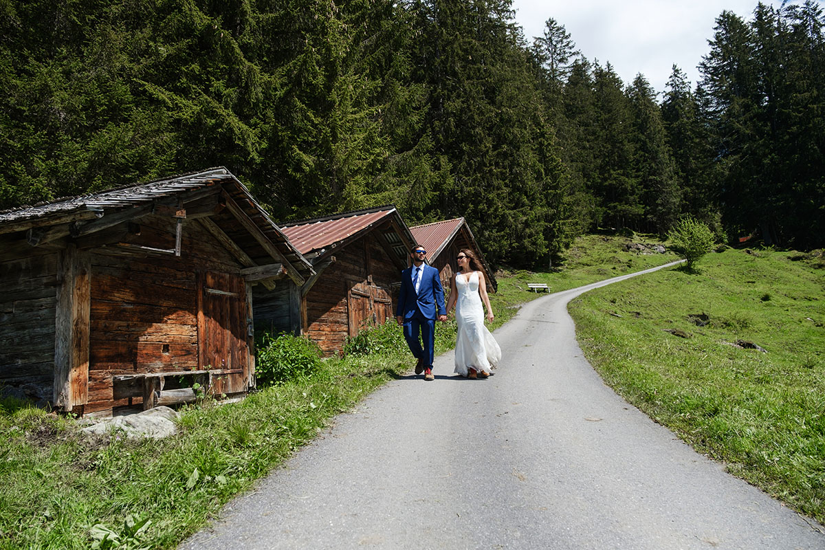 A bride and groom wearing sunglasses in bridal attire walk hand in hand past some swiss chalets during their elopement in Wengen, Switzerland