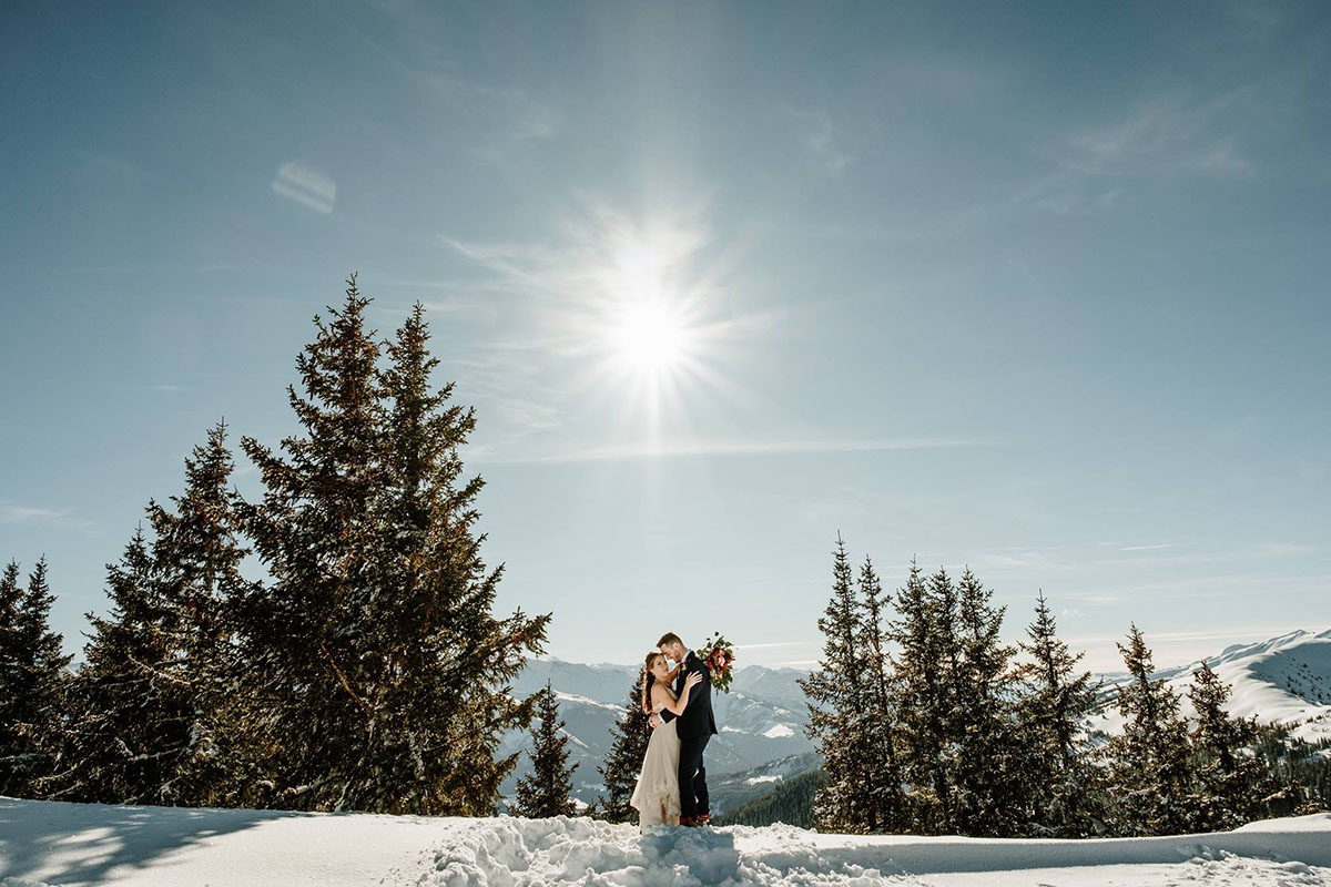 A couple in wedding clothes standing in the show embrace with the sun and trees behind them during their adventure elopement