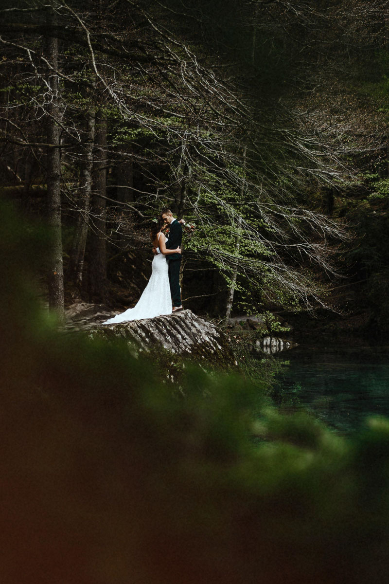 A couple stand surrounded by trees on the lakeside during their elopement in Switzerland
