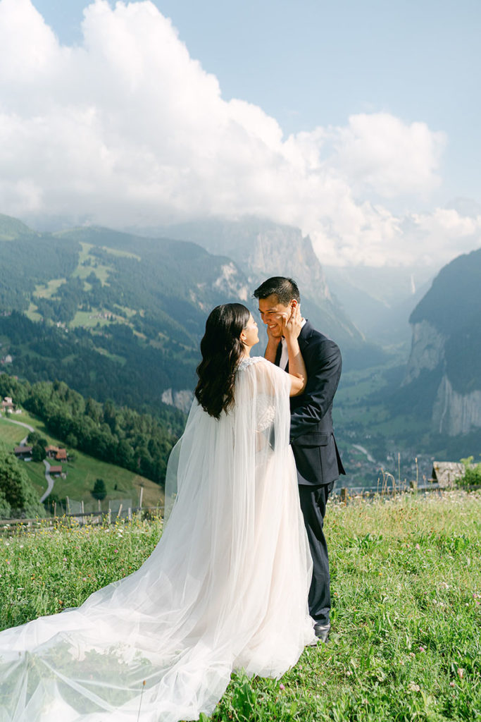 A bride in a long veil holds her grooms face above the village of Wengen during their Swiss elopement ceremony